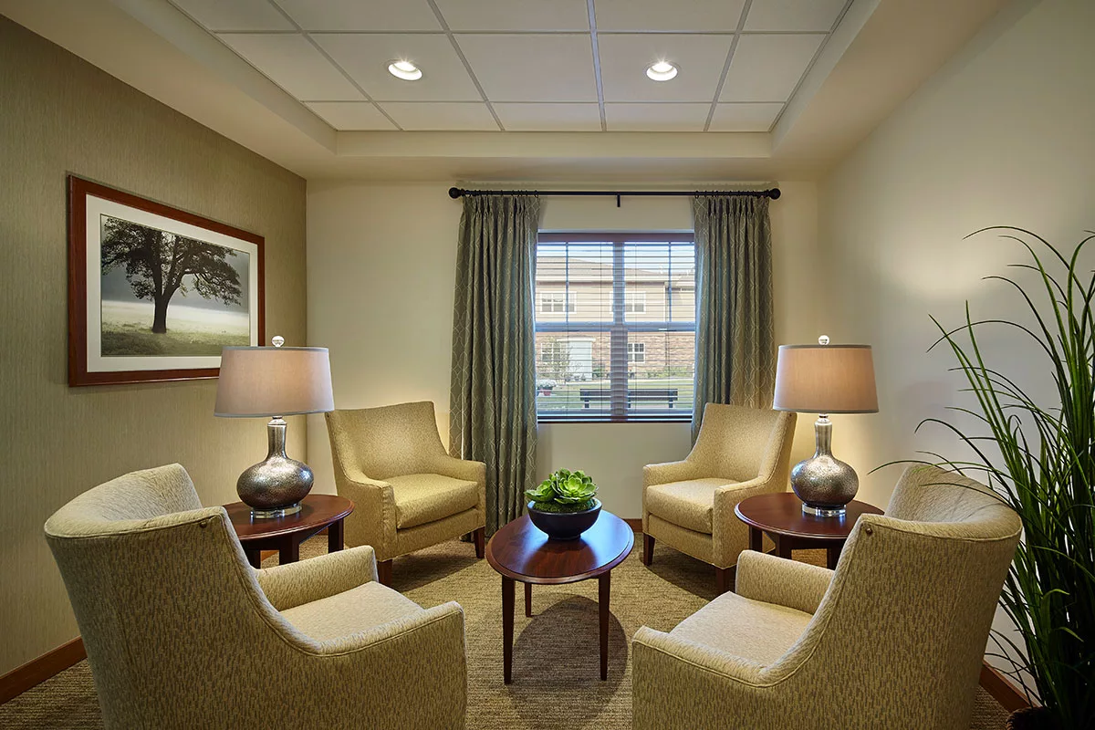 A cozy waiting room with four armchairs, two lamps, and a small central table beneath a window with curtains.