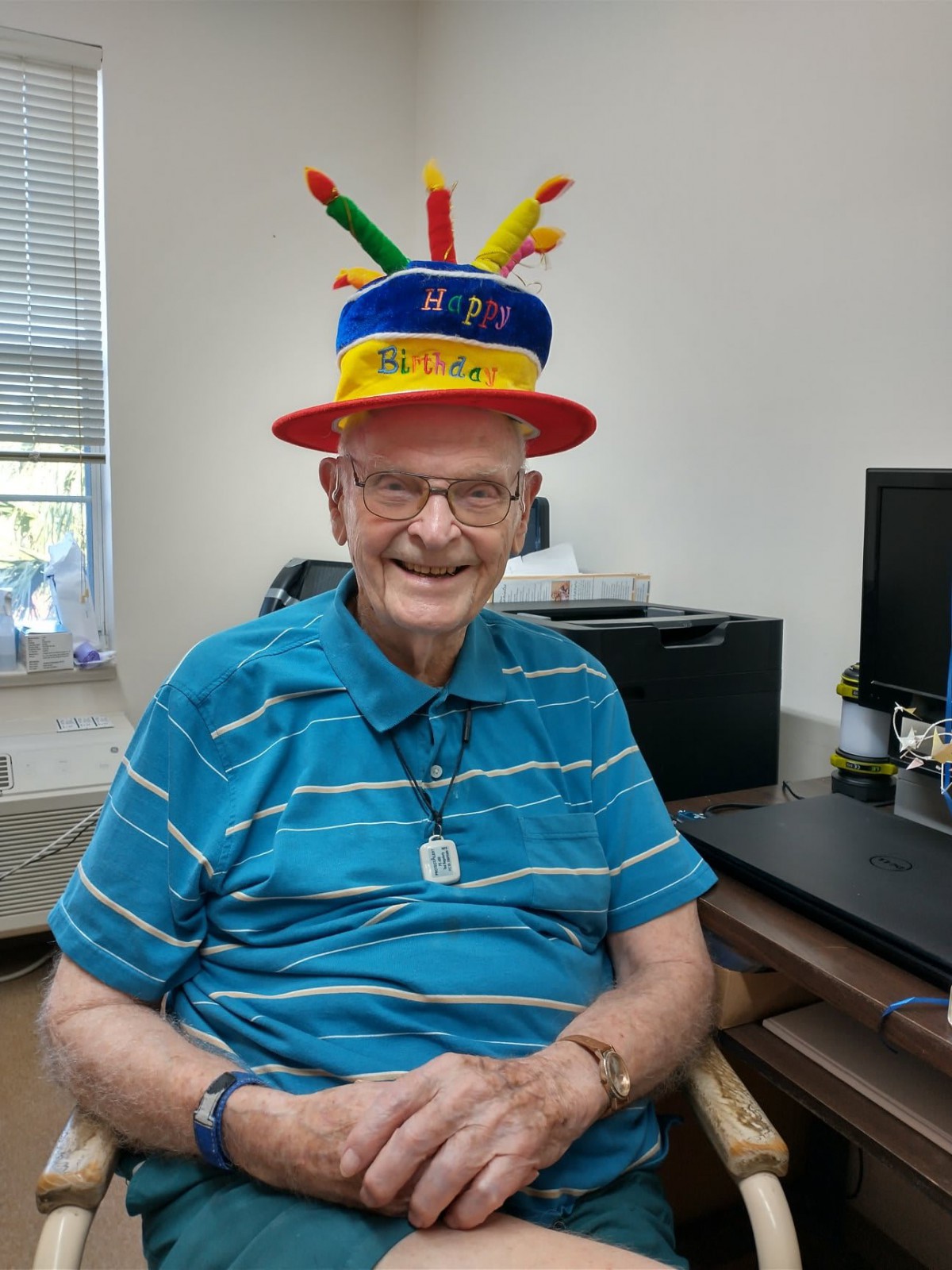 An elderly man wearing a colorful birthday hat, smiling while sitting indoors.