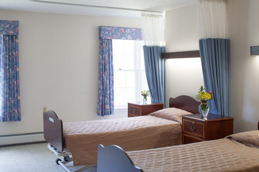 A hospital room with two patient beds, a window with blue curtains, and wooden furniture adorned with flowers.