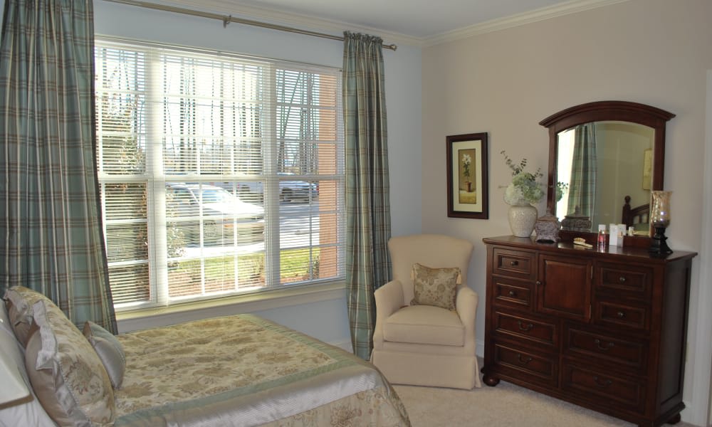 A cozy bedroom with a large window, an upholstered chair, and a wooden dresser.