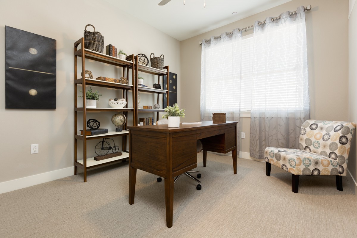 A cozy home office with a wooden desk and a bookshelf, filled with decor items, illuminated by natural light from a large window.