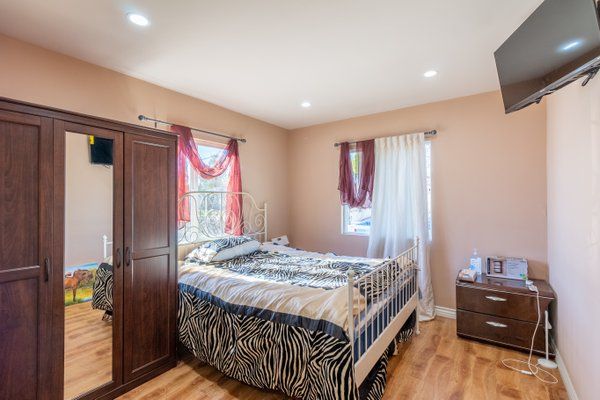 A cozy bedroom with a bed featuring zebra-patterned bedding, a wooden wardrobe, hardwood floors, and curtains on two windows.