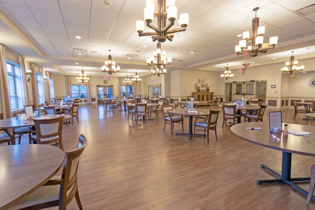 A spacious dining hall with wooden floors, numerous tables and chairs, and elegant lighting fixtures.