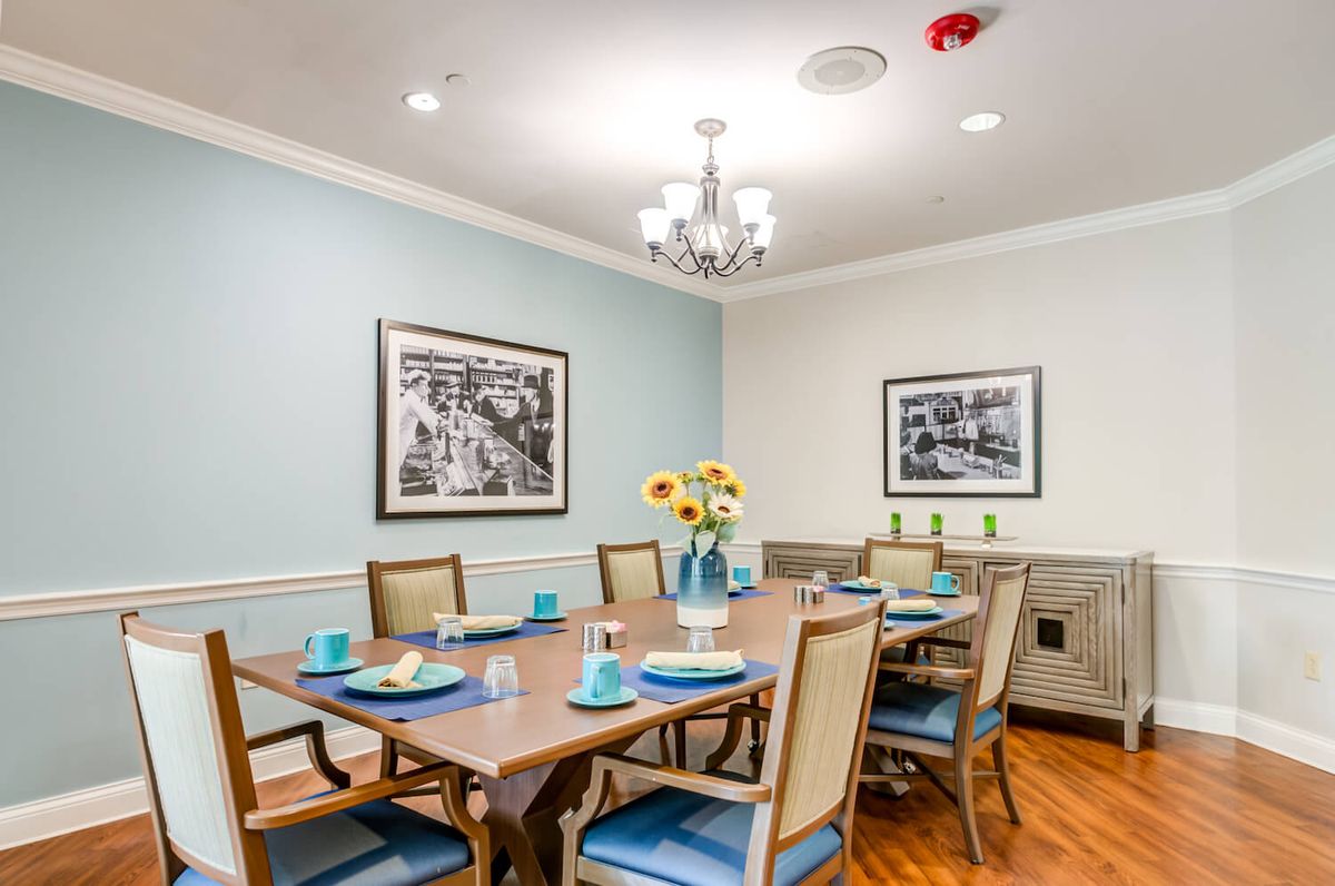 A well-appointed dining room featuring a table set for six, with blue-accented place settings and a vase of sunflowers at the center. The walls are adorned with artwork and the room is lit by a chandelier.