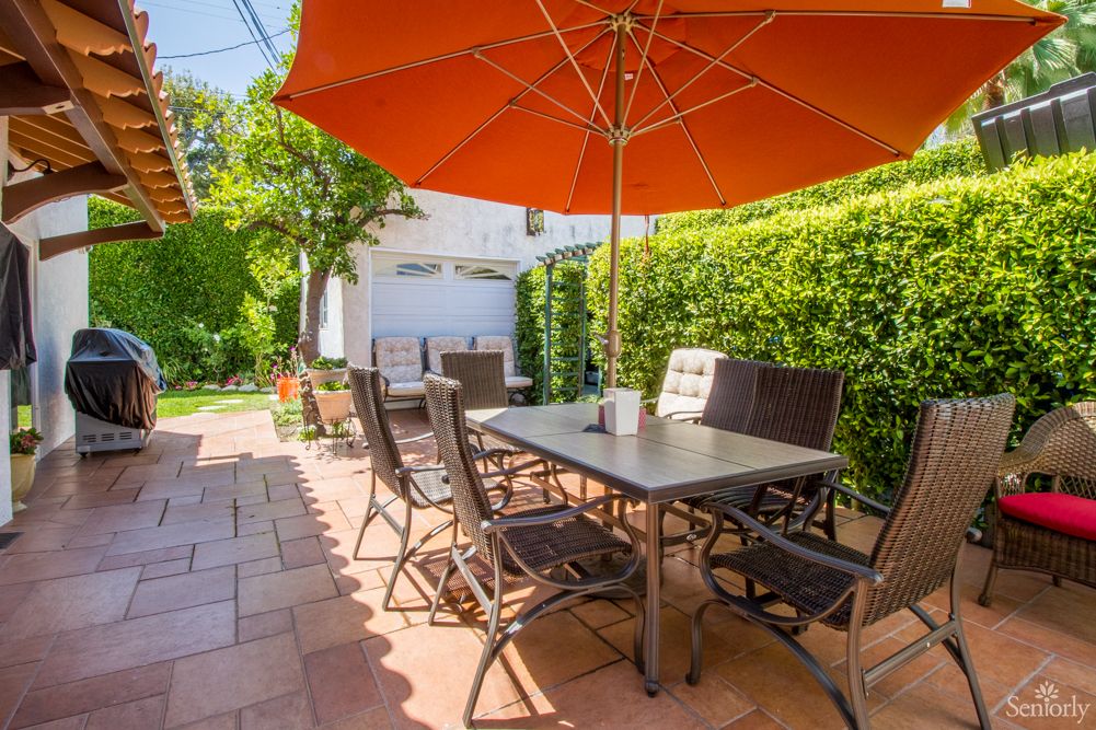 A sunny backyard patio with a dining table, chairs, and an orange umbrella.