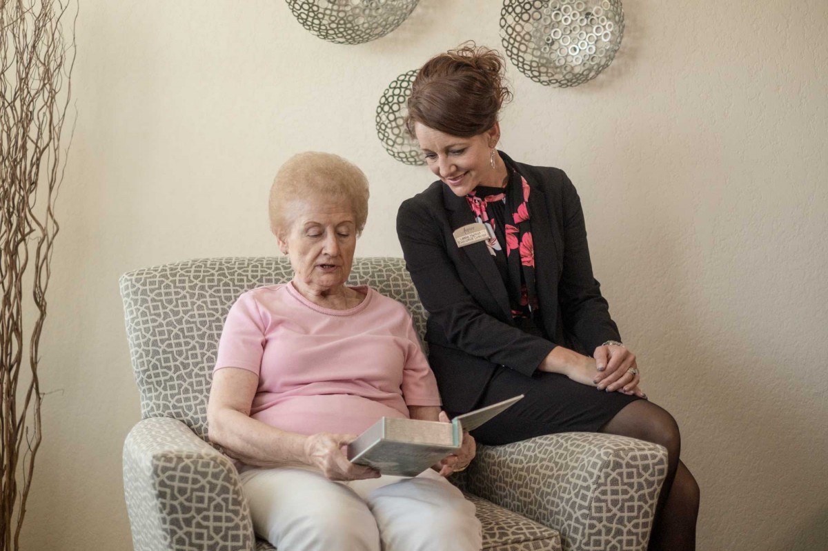 An elderly woman is reading a book while a caretaker sits beside her, engaging in conversation.