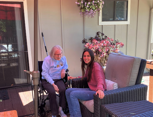 A young woman shares a moment with an elderly person in a wheelchair, surrounded by flowers on a sunny day.