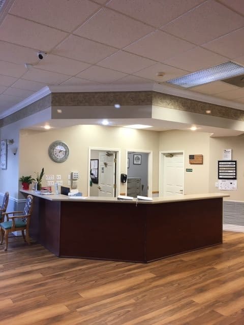 A spacious reception area with a central desk and waiting chairs, featuring a clock and well-lit ceiling.