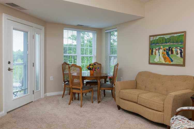 A cozy living room with a dining area featuring a round table and chairs, a beige couch, and large windows bringing in natural light.