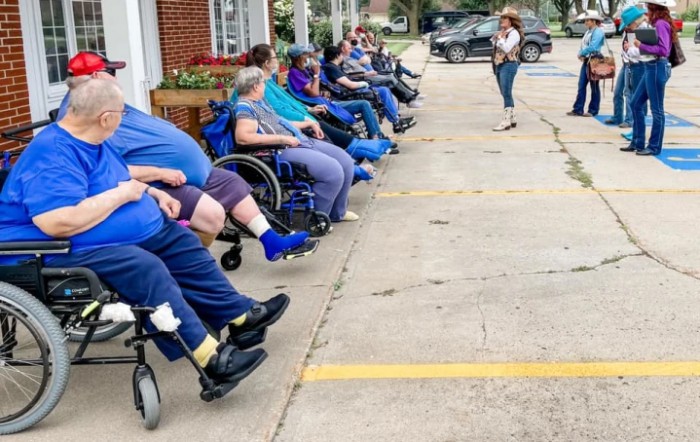 A group of people seated in wheelchairs outdoors, listening to a person speaking in a parking lot.