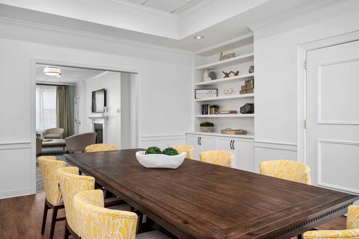 A modern dining room with a long wooden table and yellow chairs, featuring built-in shelves with decorative items.