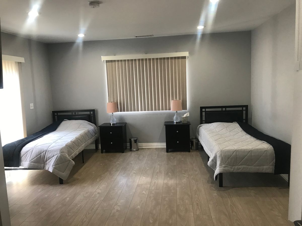 A minimalist bedroom with two twin beds, each accompanied by a nightstand and lamp, under window blinds.