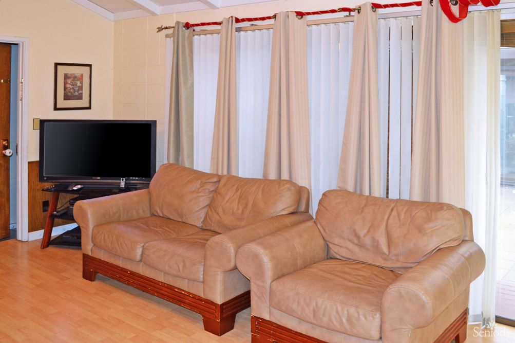 A cozy living room with two beige sofas, a television in the corner, and light-colored curtains covering the windows.