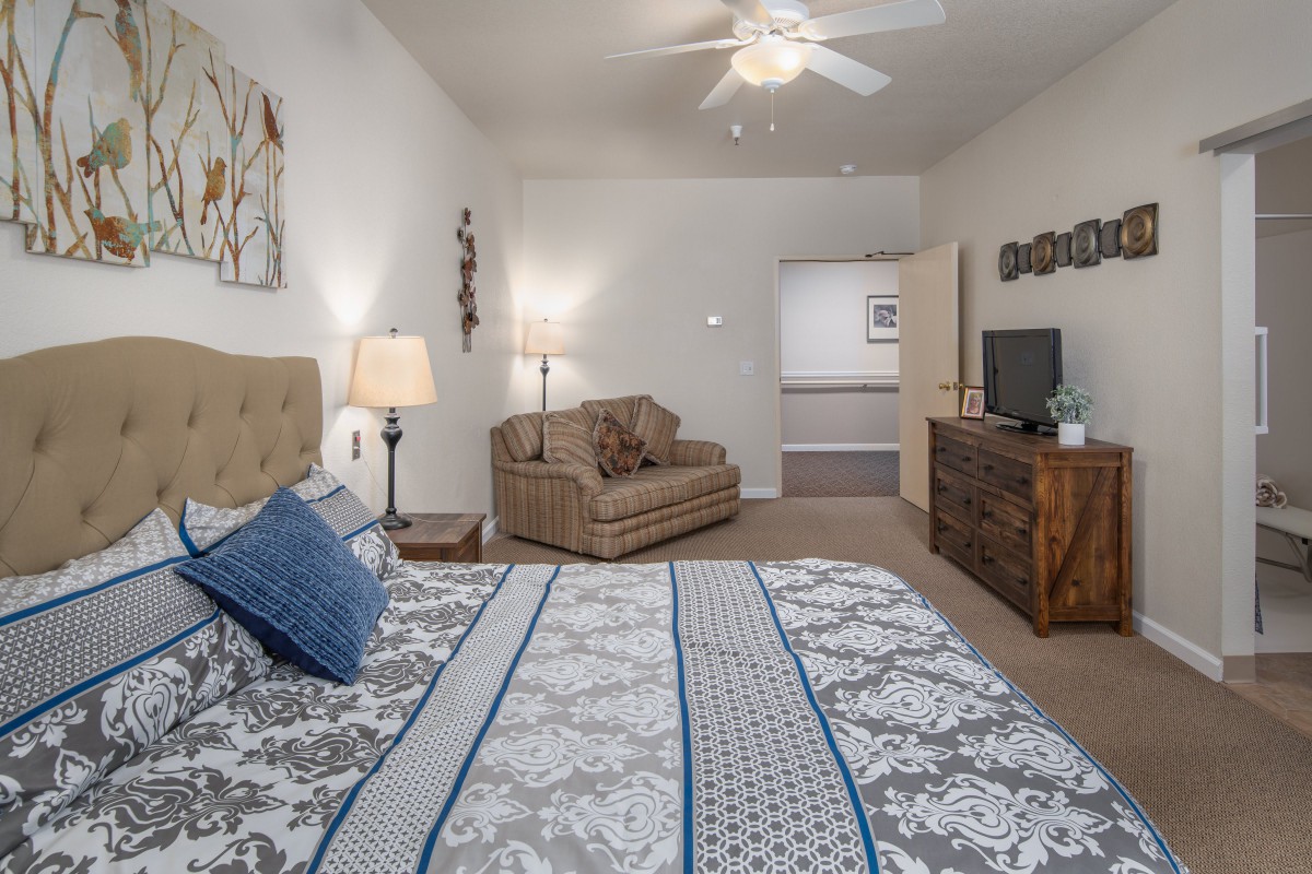A cozy bedroom featuring a comfortable bed, a sofa chair, a TV on a dresser, and warm lighting.