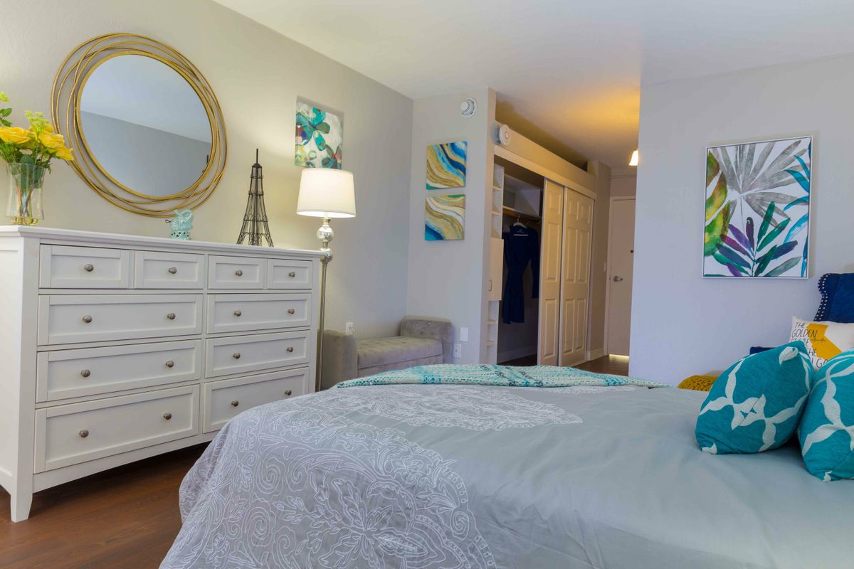 A cozy bedroom with a neatly made bed, a dresser topped with a lamp and mirror, and decorative artwork on the walls.