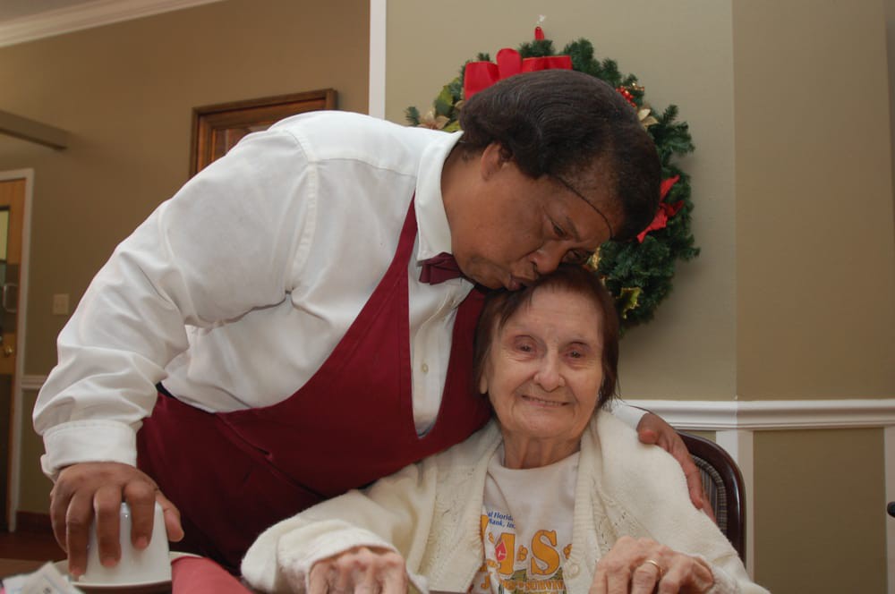 A caregiver shows affection to an elderly woman in a dining setting.