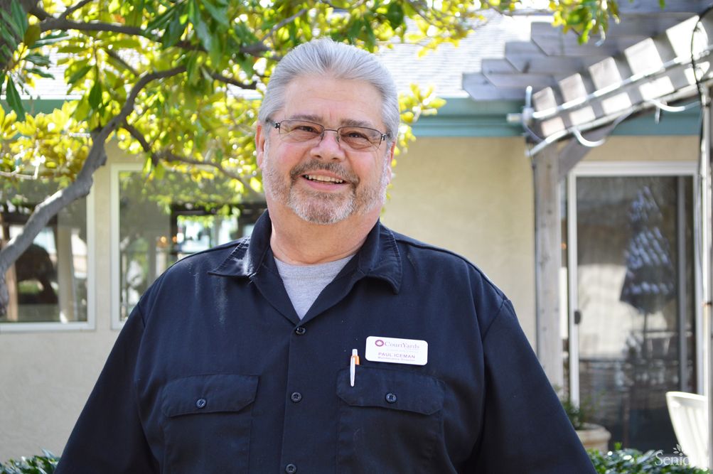 A man smiling outdoors, wearing a name tag and casual clothing.