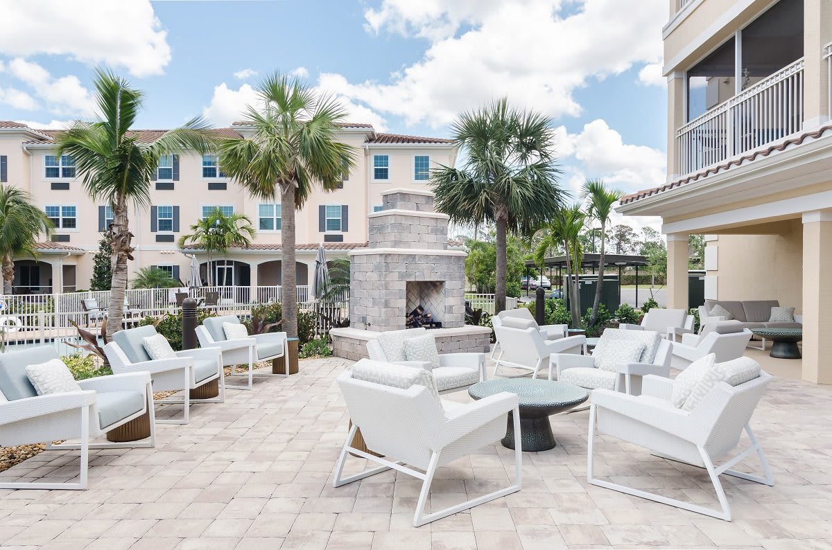 A modern outdoor patio with white furniture and a stone fireplace, surrounded by palm trees and adjacent buildings.