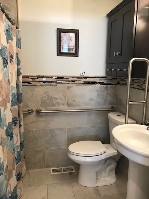 A small bathroom with a toilet, sink, and shower curtain. The walls are tiled and there is a grab bar next to the toilet.