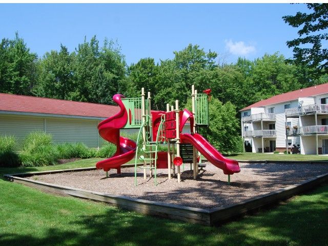 A sunny playground featuring red slides surrounded by green grass and trees.