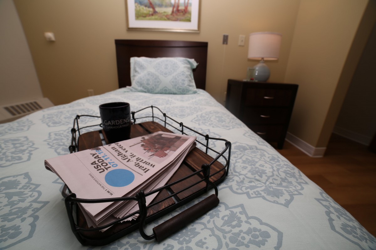 A cozy bedroom scene with a bed, bedside lamp, and tray with newspapers.