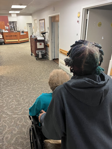 A caregiver pushing an elderly person in a wheelchair through a healthcare facility.