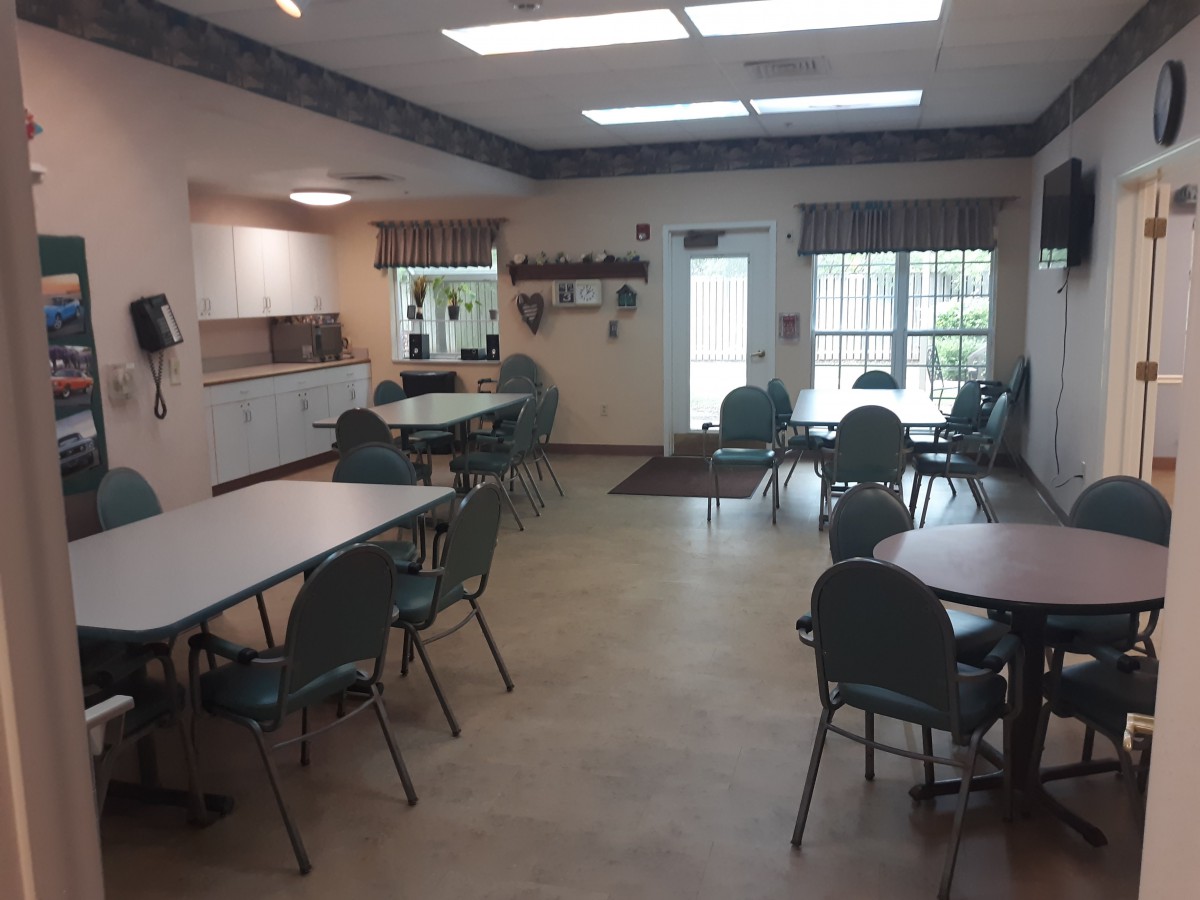 A spacious community center meeting room with tables and chairs, featuring a kitchen area and large windows.