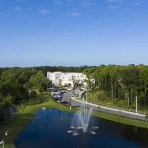 Aerial view of a building surrounded by trees and a pond with a fountain under a clear blue sky.