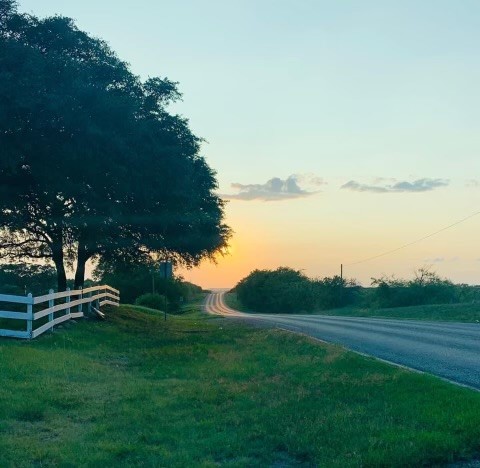 A serene sunset view along a peaceful road, framed by lush greenery and a white fence.