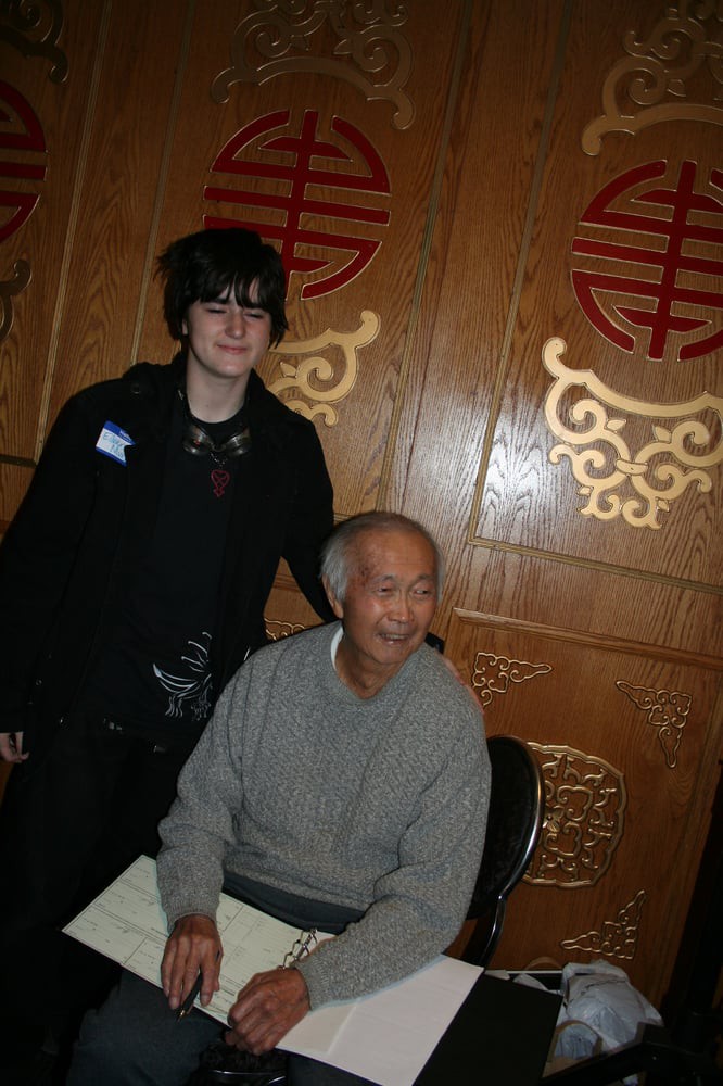 A young person standing next to an older seated individual in front of a wooden panel with ornate designs.