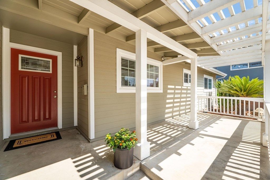 A sunny front porch with a red door, potted plants, and wooden columns casting shadows.