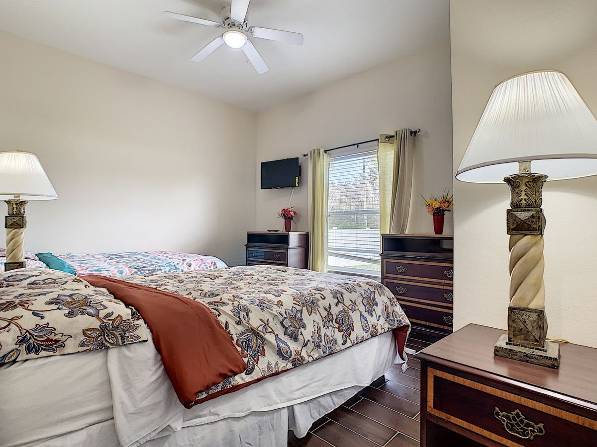 A cozy bedroom featuring a double bed with floral bedding, a nightstand with a lamp, and a dresser next to a window with curtains.