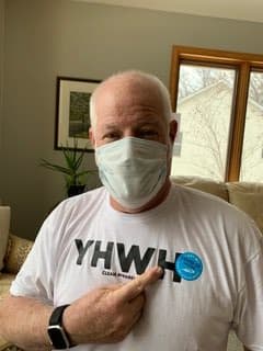 A person wearing a mask and a t-shirt with 'YHWH' printed on it, pointing to a sticker while indoors.