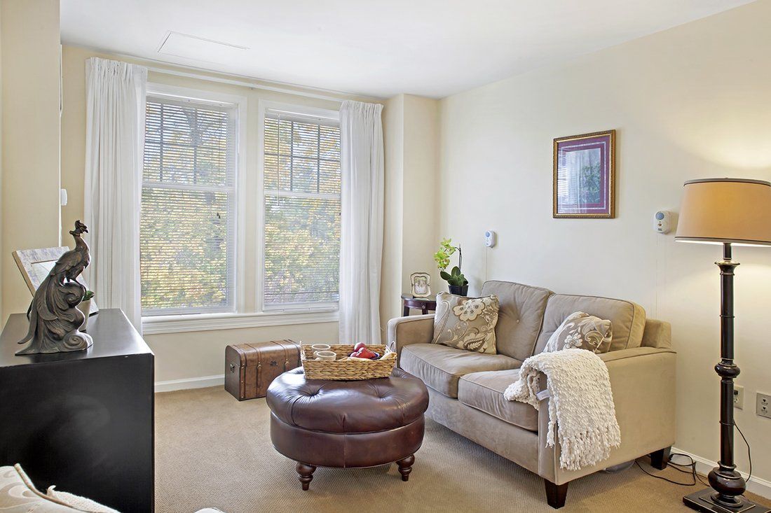 A cozy living room with a sofa and ottoman, illuminated by natural light from large windows.