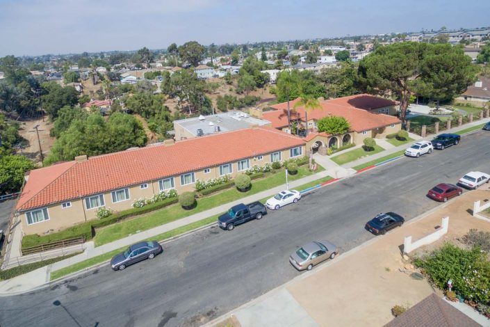 An aerial view of a suburban residential area with cars parked along the street and a long building with a red-tiled roof.