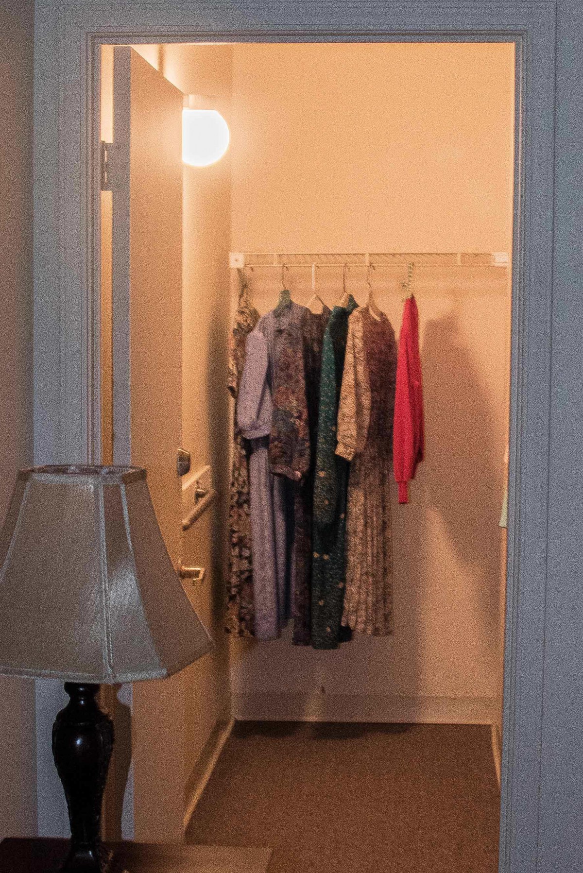 A small closet with clothes hanging and a nearby lamp illuminating the space.
