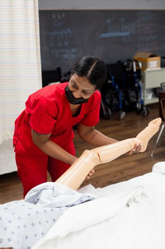 A nursing student in red scrubs practices patient care techniques using a mannequin leg.