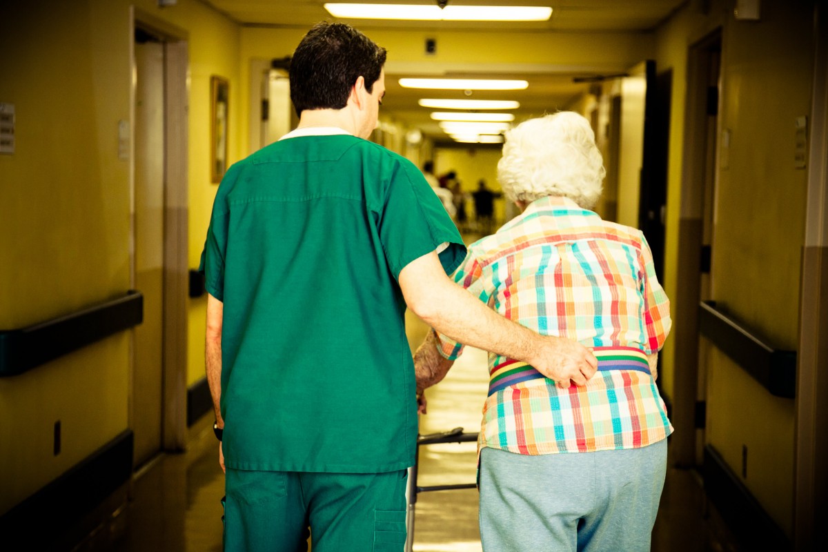 A healthcare worker assists an elderly person down a hospital corridor.