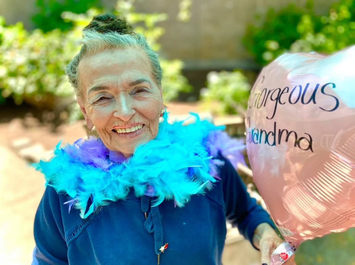 An elderly woman smiling and holding a balloon, wearing a colorful feather boa outdoors.