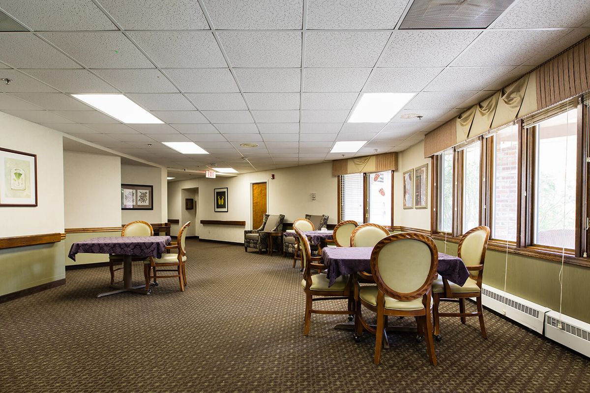 A spacious dining room with tables and chairs arranged neatly, featuring carpeted flooring and large windows allowing natural light.