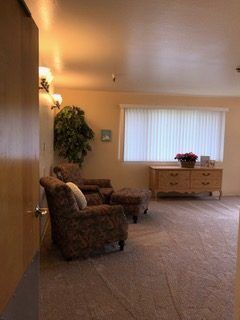 A cozy living room featuring floral armchairs, a wooden dresser, and a large window with vertical blinds.