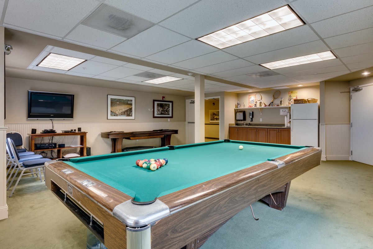 A well-lit recreation room featuring a pool table and a small kitchenette in the background.