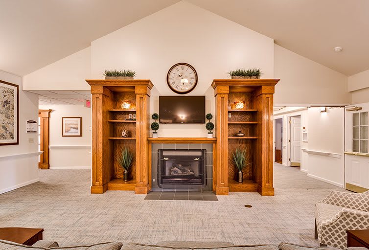 A cozy living room setting featuring a fireplace flanked by wooden shelves, with a TV and clock above.