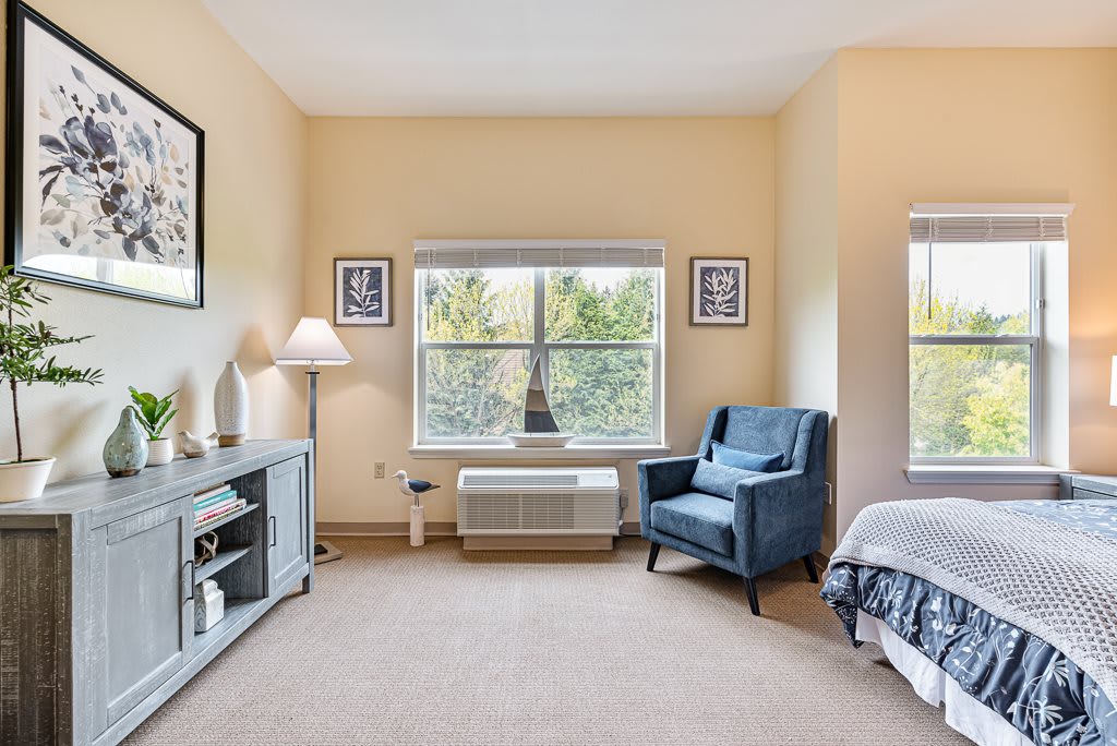 A cozy bedroom with a large window, featuring an armchair, a lamp, and decorative elements.