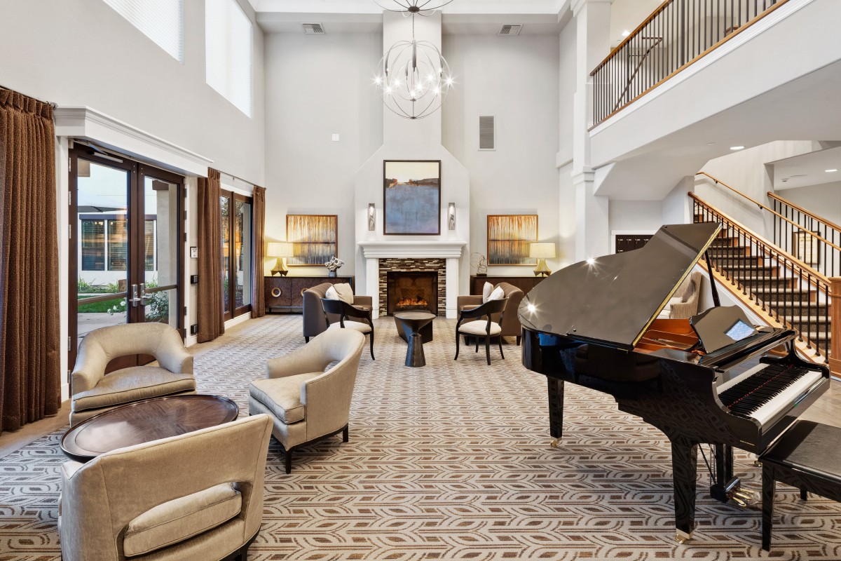 A luxurious living room featuring a grand piano, elegant furniture, and a fireplace under a high ceiling with a chandelier.