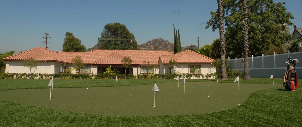 A house with a putting green in the foreground, surrounded by trees and mountains.