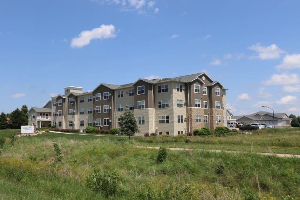 A multi-story apartment building surrounded by a grassy field under a blue sky with clouds.