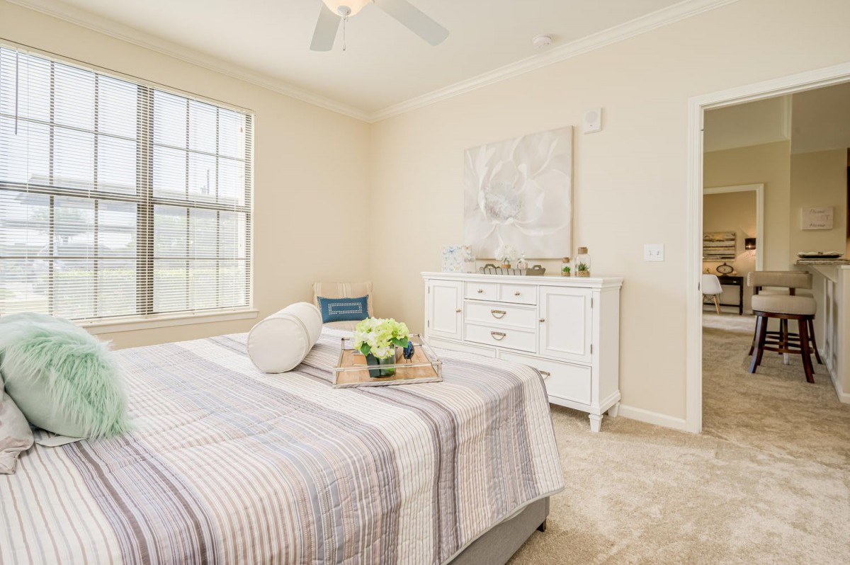 A cozy bedroom with neutral tones featuring a made bed, a dresser, and large windows.
