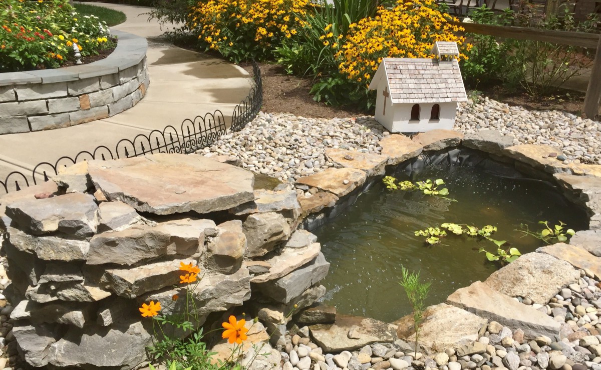 A serene garden setting featuring a pond with water lilies, surrounded by rocks and vibrant flowers, along with a decorative miniature house.