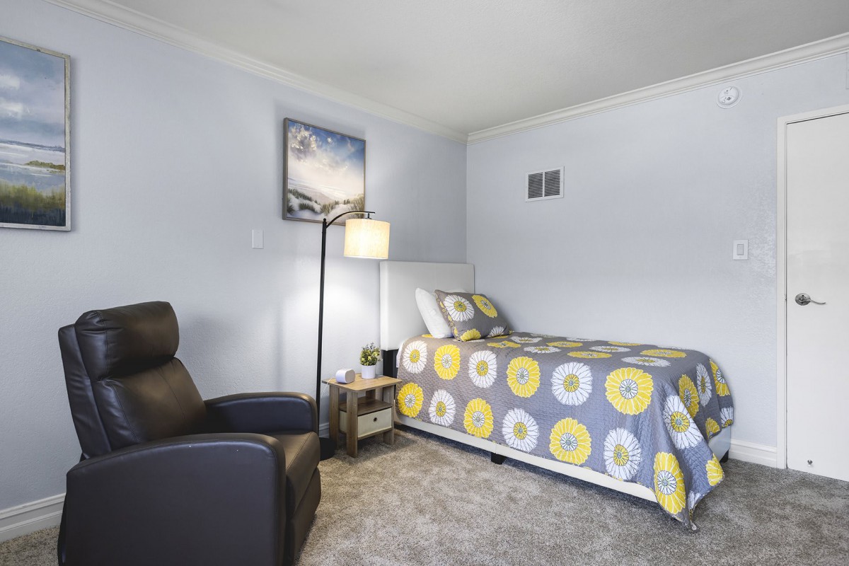 A cozy bedroom with a single bed featuring a colorful duvet, an armchair, and a standing lamp.